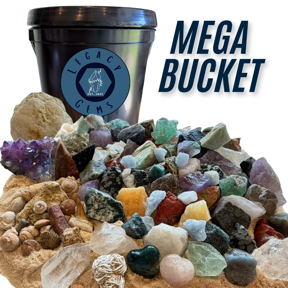 A large assortment of raw gemstones, crystals, fossils, and small rocks displayed in front of a black plastic gallon bucket with a blue Legacy Gems logo. Large text on the right reads 'MEGA BUCKET'.
