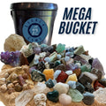 A large assortment of raw gemstones, crystals, fossils, and small rocks displayed in front of a black plastic gallon bucket with a blue Legacy Gems logo. Large text on the right reads 'MEGA BUCKET'.