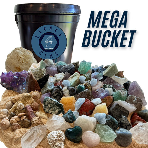 A large assortment of raw gemstones, crystals, fossils, and small rocks displayed in front of a black plastic gallon bucket with a blue Legacy Gems logo. Large text on the right reads 'MEGA BUCKET'.