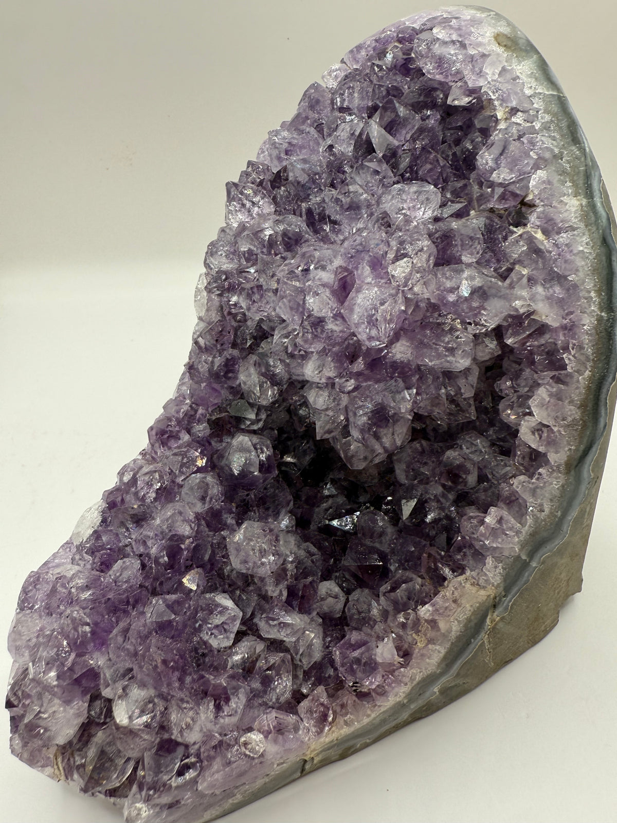 A natural amethyst crystal geode specimen with dense purple and lavender crystal clusters inside, set in a gray stone matrix on a plain light background.