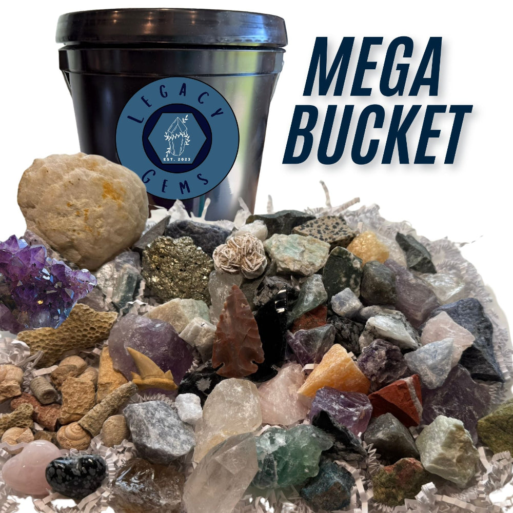 A large assortment of raw gemstones, crystals, and small rocks piled in front of a black bucket with a blue Legacy Gems logo. Large text on the right reads 'MEGA BUCKET'.